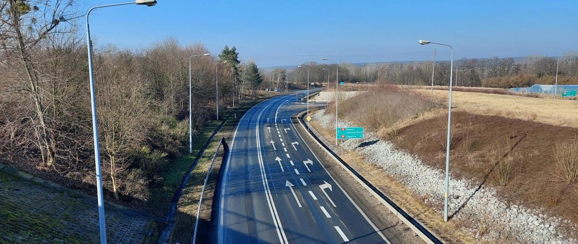 Fotografia przedstawia widok z góry na drogę w słoneczny dzień. Na jezdni widoczne są strzałki kierunkowe, wyznaczające pasy ruchu. Po obu stronach drogi znajdują się latarnie uliczne. Lewa strona jest obsadzona drzewami pozbawionymi liści, natomiast po prawej stronie widoczne są suche trawy i krzewy. W oddali znajduje się zielony drogowskaz z białymi napisami, wskazujący kierunki do miast: „A4 Wrocław”, „Zielona Góra” oraz „Legnica”.