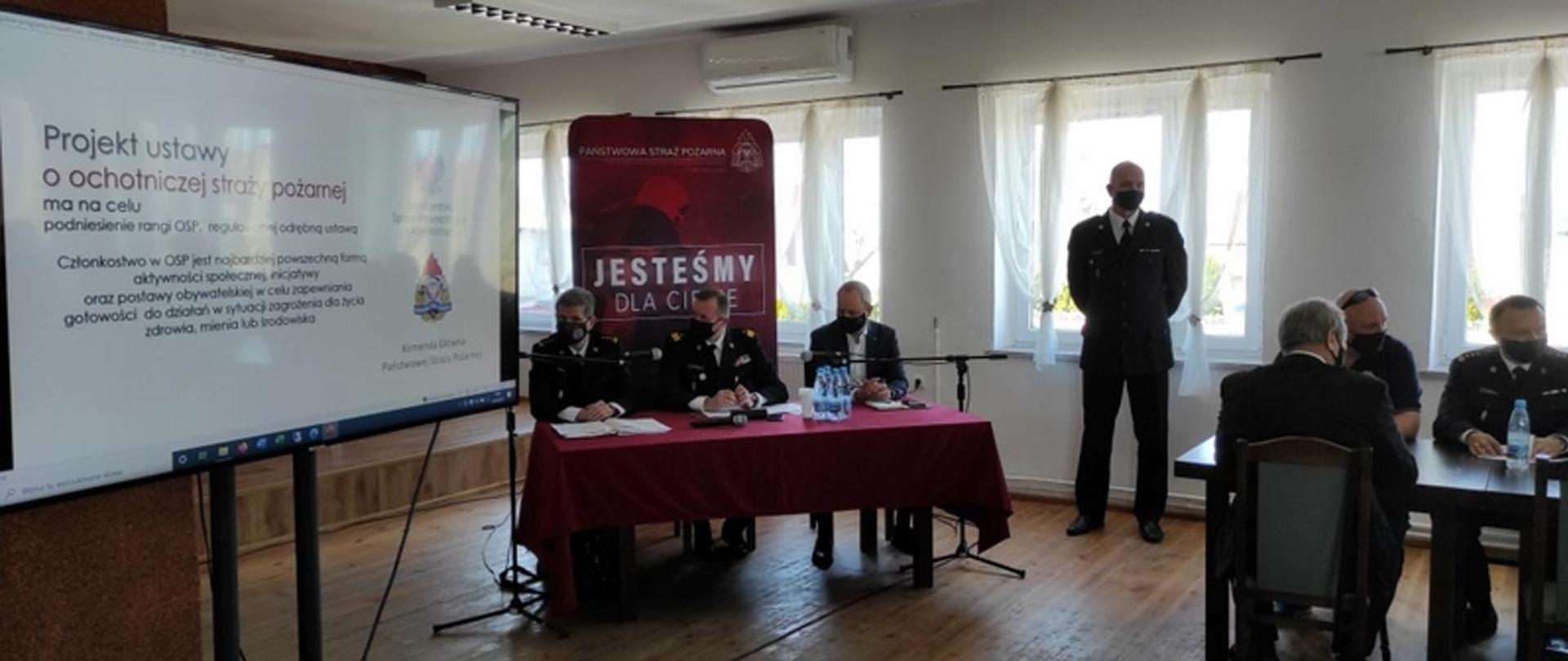Po lewo wyświetlana jest prezentacja w zakresie konsultacji projektu ustawy, na wprost siedzi komendant wojewódzki PSP, zastępca komendanta głównego PSP, poseł na sejm RP, po prawej stronie przy stole siedzą inni uczestnicy konsultacji