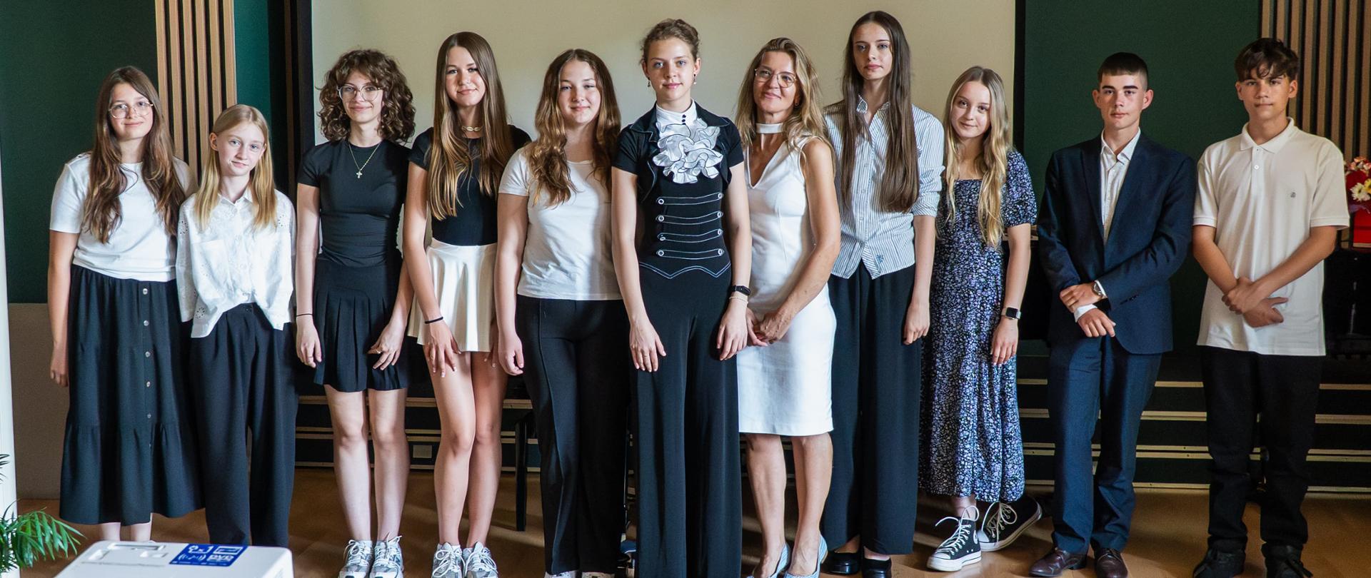 Na zdjęciu widzimy grupę uczniów, którzy pozują do wspólnego zdjęcia podczas uroczystości zakończenia roku szkolnego. Wszyscy są elegancko ubrani, co podkreśla uroczysty charakter wydarzenia. W centrum stoi kilka dziewcząt w białych i ciemnych strojach, a obok nich znajduje się chłopak w garniturze. Wszyscy wyglądają na radosnych i dumnych, co wskazuje na znaczenie tego momentu w ich życiu. W tle widoczny jest fortepian oraz projektor, co sugeruje, że wydarzenie może obejmować różne wystąpienia lub prezentacje. Atmosfera jest pełna pozytywnych emocji i entuzjazmu.