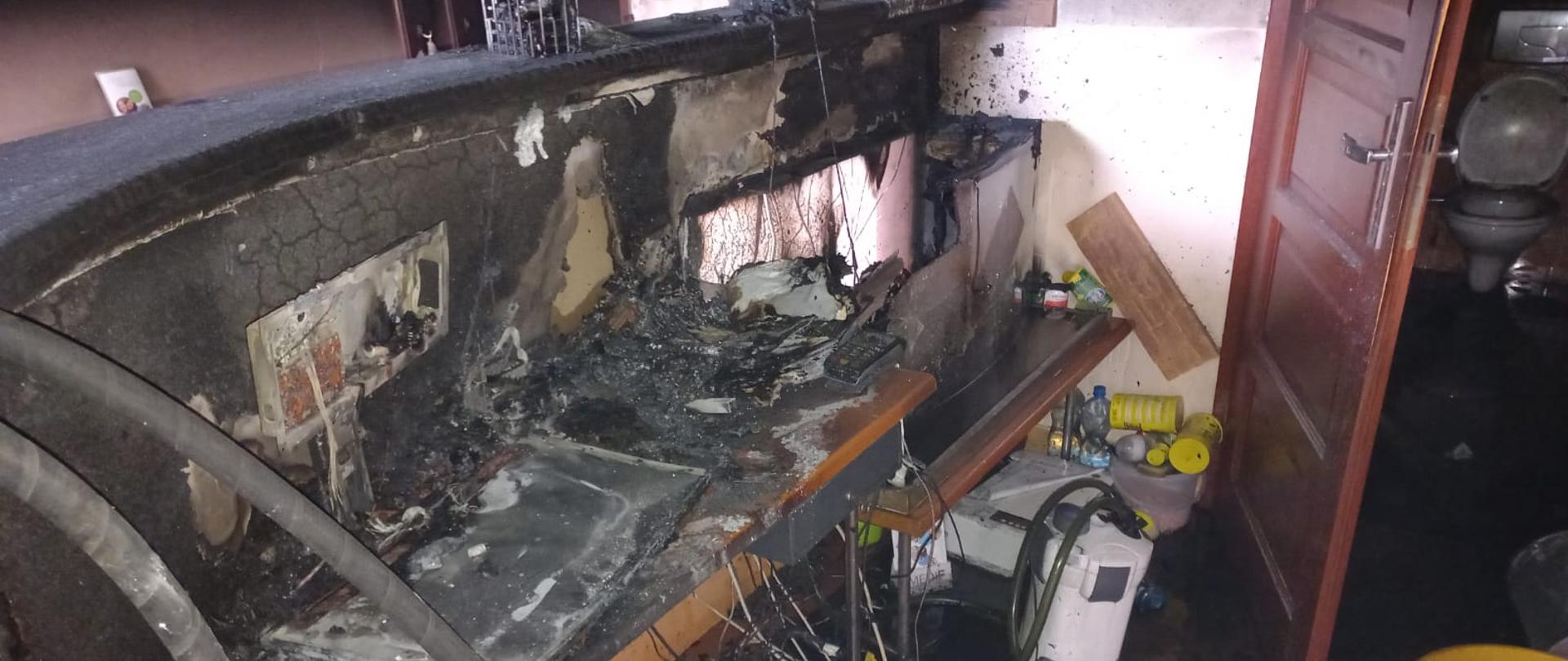 Widok spalonej lady recepcyjnej wraz ze znajdującymi się na niej urządzeniami. Na ścianach widoczne znaczne okopcenia.