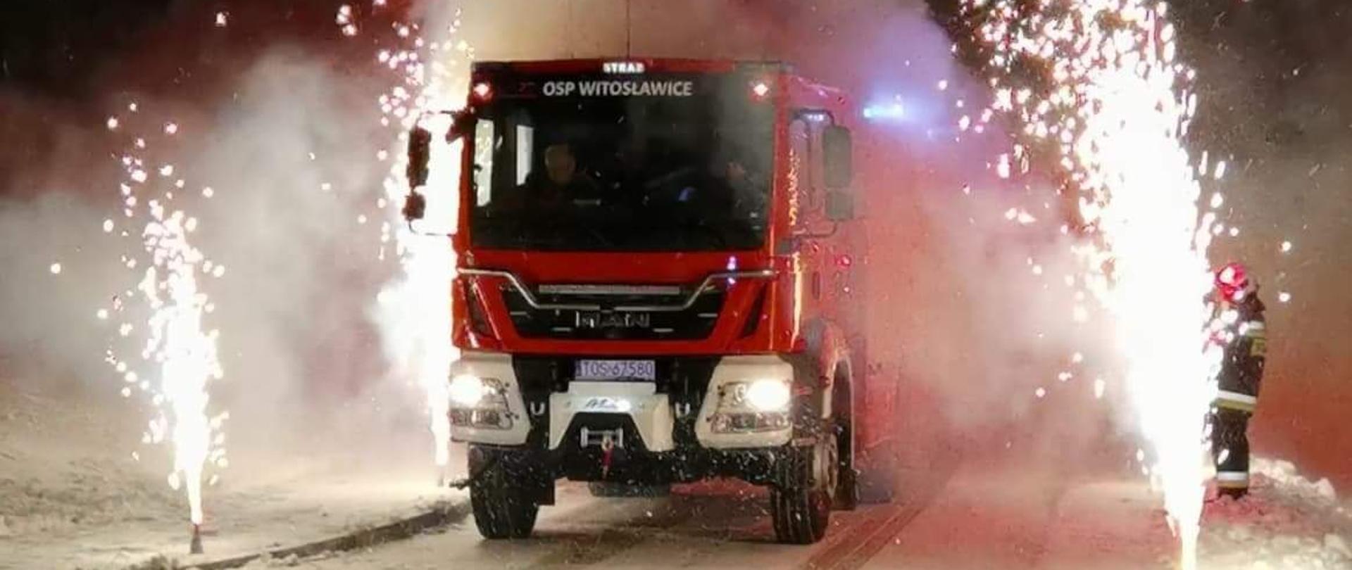 Na pierwszym planie w porze nocnej czerwony samochód gaśniczy podczas przyjęcia do jednostki. Wokół teren rozświetlony białymi racami. Po prawej stronie w tle stoi umundurowany strażak.