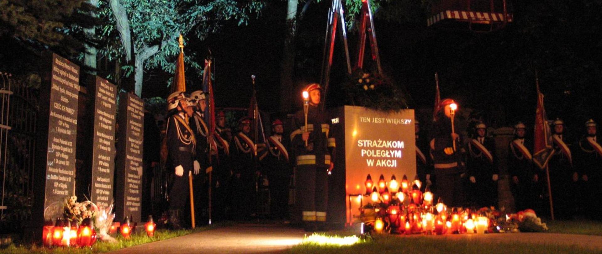 Zdjęcie przedstawia Strażaków w mundurach galowych i bojowych przed pomnikiem upamiętniającym poległych strażaków. Przed pomnikiem ustawione znicze.