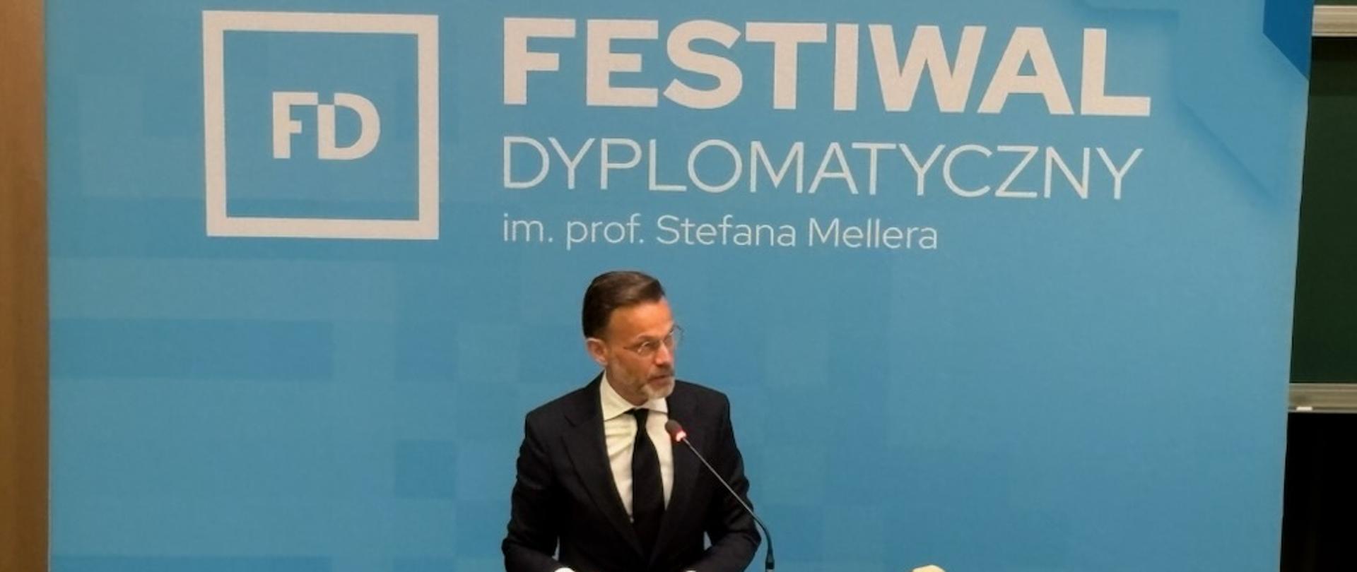 Mężczyzna w garniturze stoi przy mównicy, wygłaszając przemówienie. 