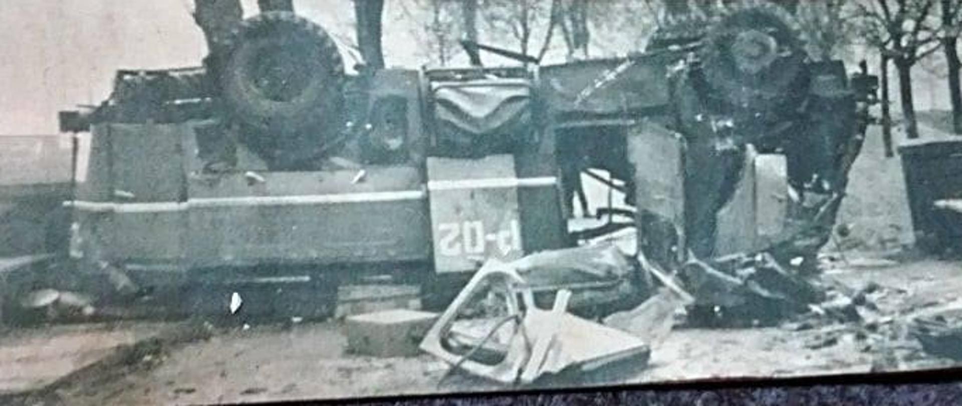 Zdjęcie z wypadku przedstawiające rozbity samochód pożarniczy znajdujący się w poprzek drogi, w pozycji kołami do góry. 