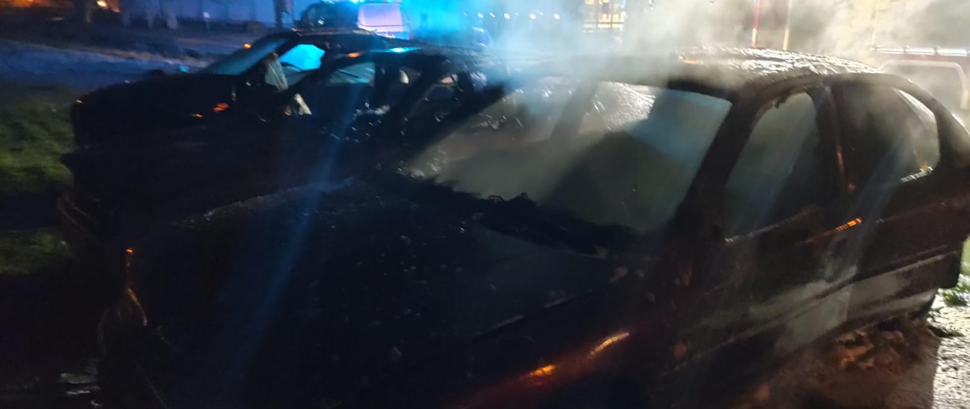 Na zdjęciu trzy uszkodzone samochody, pierwszy częściowo pokryty pianą gaśniczą. Nad nimi unosi się dym, w tle samochód ratowniczo-gaśniczy państwowej straży pożarnej, dalej samochód policji.