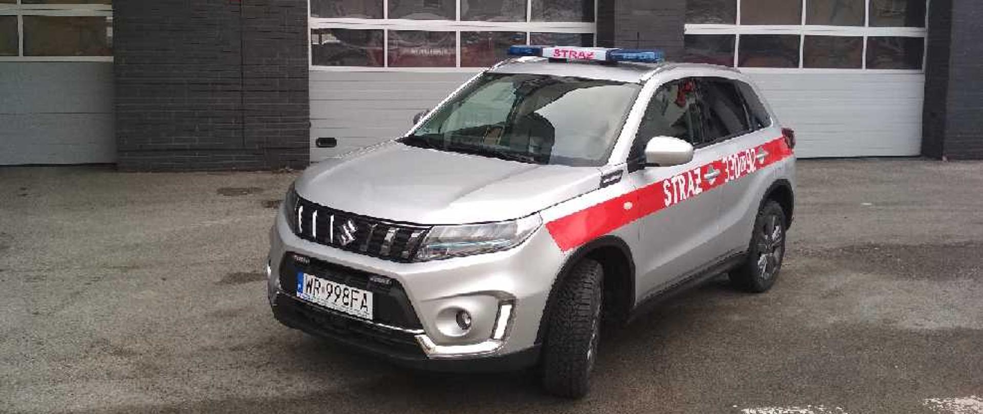 Zdjęcie przedstawia nowy lekki pojazd operacyjny marki Suzuki Vitara. Pojazd typu suv wyposażony w napęd terenowy na wszystkie cztery koła. Karoseria pojazdu pomalowana jest w kolorze szarym. Po bokach pojazdu znajduje się czerwony pas na którym umieszczone są białe cyfry i litery tworzące numer rozpoznawczy - tak zwany numer operacyjny właściwy dla danego pojazdu. Na dachu pojazdu umieszczona jest alarmowa belka sygnałowa z centralnie położonym napisem straż. Zdjęcie wykonane przed garażami obiektu Komendy Miejskiej Państwowej Straży Pożarnej w Radomiu oraz Jednostki Ratowniczo - Gaśniczej numer 1 w Radomiu. Na zdjęciu widoczny przód oraz lewy bok pojazdu.