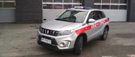 Zdjęcie przedstawia nowy lekki pojazd operacyjny marki Suzuki Vitara. Pojazd typu suv wyposażony w napęd terenowy na wszystkie cztery koła. Karoseria pojazdu pomalowana jest w kolorze szarym. Po bokach pojazdu znajduje się czerwony pas na którym umieszczone są białe cyfry i litery tworzące numer rozpoznawczy - tak zwany numer operacyjny właściwy dla danego pojazdu. Na dachu pojazdu umieszczona jest alarmowa belka sygnałowa z centralnie położonym napisem straż. Zdjęcie wykonane przed garażami obiektu Komendy Miejskiej Państwowej Straży Pożarnej w Radomiu oraz Jednostki Ratowniczo - Gaśniczej numer 1 w Radomiu. Na zdjęciu widoczny przód oraz lewy bok pojazdu.