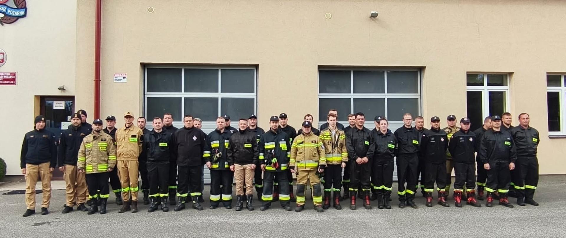 Duża grupa kilkudziesięciu strażaków OSP i PSP w umundurowaniu (ubrania specjalne i mundury służbowe w kolorach czarnym, beżowym i jasnożółtym) stoi w szeregu na placu przed budynkiem strażnicy. Zdjęcie grupowe absolwentów szkolenia kierowców-konserwatorów sprzętu ratowniczego OSP.