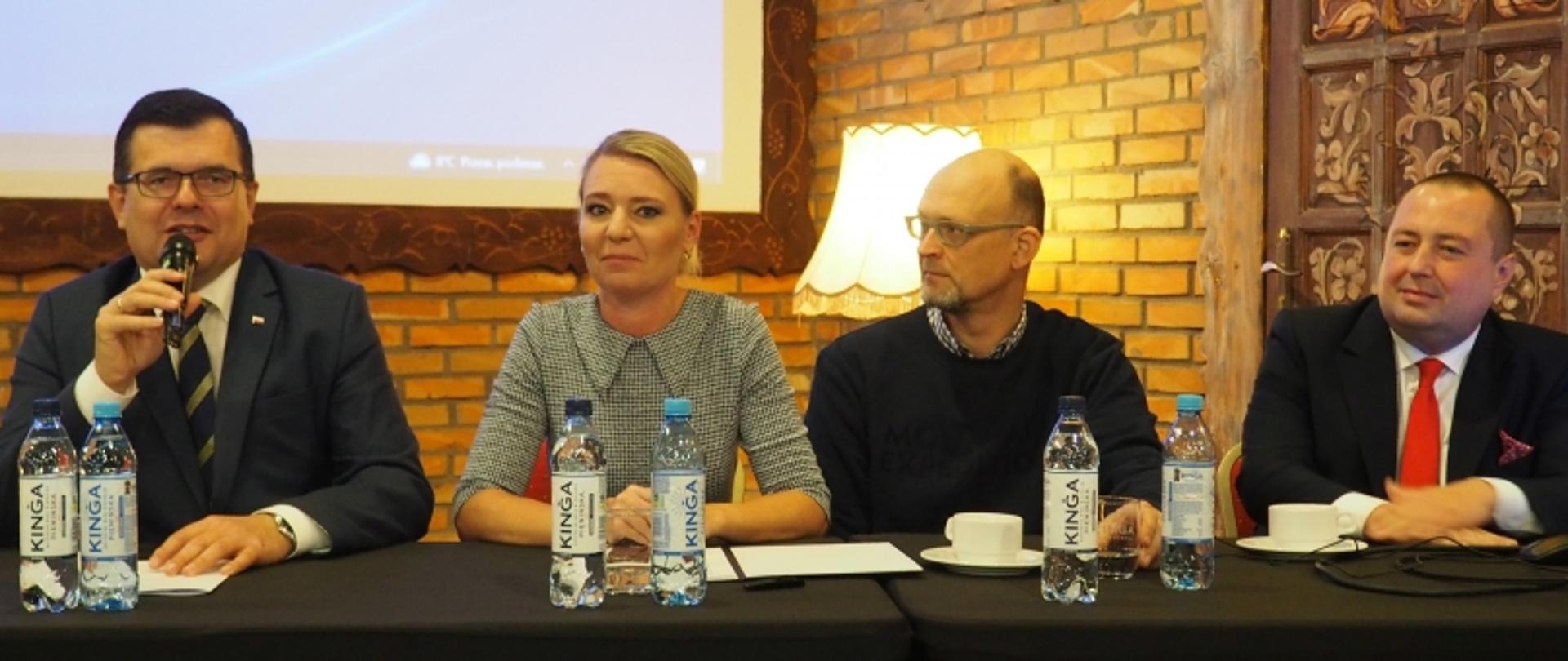 Dwie osoby stojące przed banerem GUNB i drewnianą szafą, trzymające granatowe teczki z godłem.