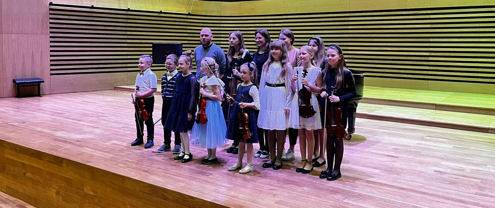 W dwóch rzędach na estradzie stoi dziesięcioro dzieci, dwie kobiety oraz mężczyzna.