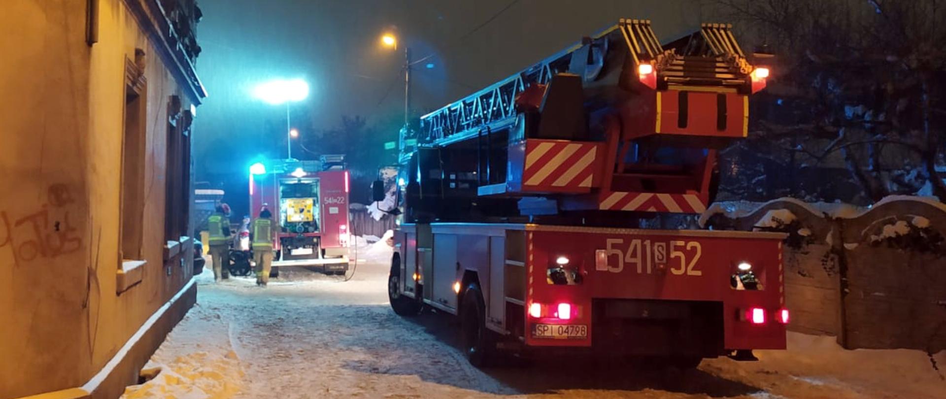 Zdjęcie przedstawia z lewej strony widoczny jest fragment budynku mieszkalnego, z prawej strony widoczny jest samochód specjalny - drabina mechaniczna. W tle widoczny samochód ratowniczo - gaśniczy oraz strażacy w ubraniach specjalnych wykonujący czynności ratownicze.