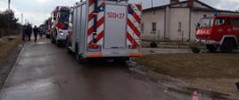 Zdjęcie zrobione na drodze gminnej, ulica Słoneczna miejscowość Mzyki. Wzdłuż drogi ustawione samochody pożarnicze. Droga zablokowana za pomocą ustawionych pachołków ostrzegawczych.