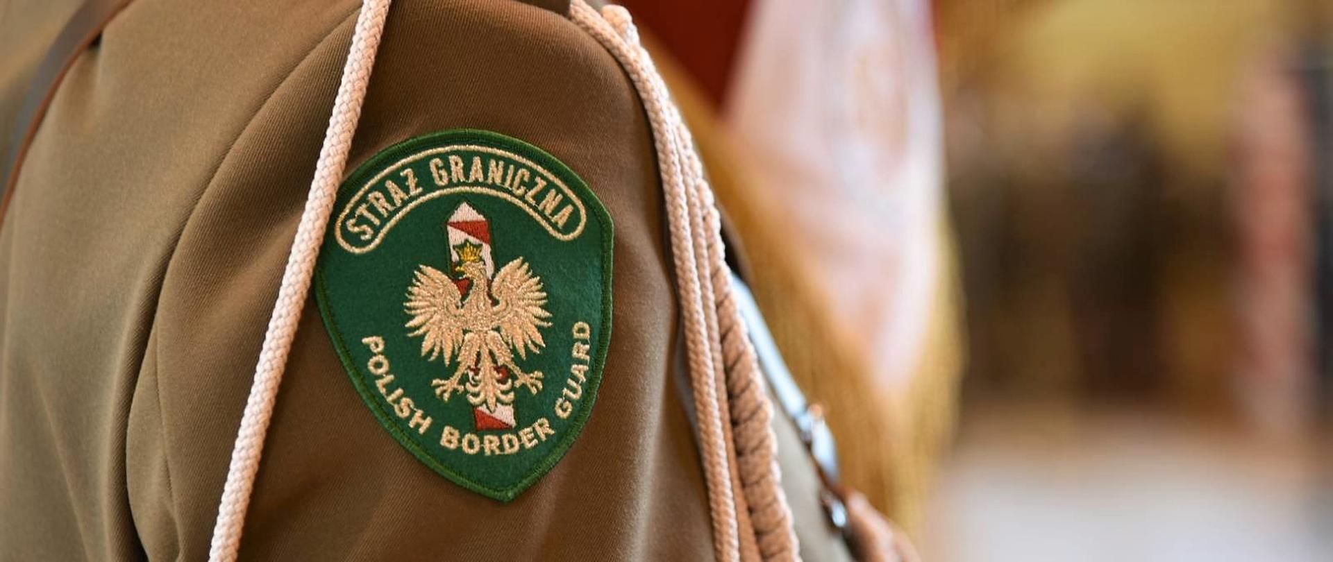 Zielona naszywka na rękawie munduru z napisem "Straż Graniczna Polish Border Guard" okalający orła białego w złotej koronie umieszczonego na tle biało-czerwonego słupka granicznego 