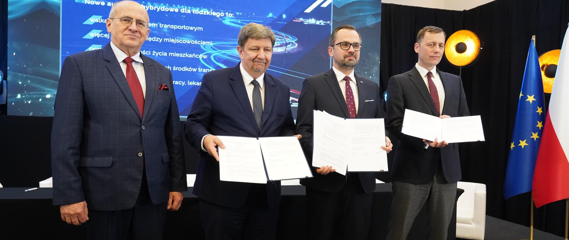 Cztery osoby stoją obok siebie. Drugi od prawej stoi wiceminister Marcin Horała. Trzech mężczyzn trzyma dokumenty w obu dłoniach. 
