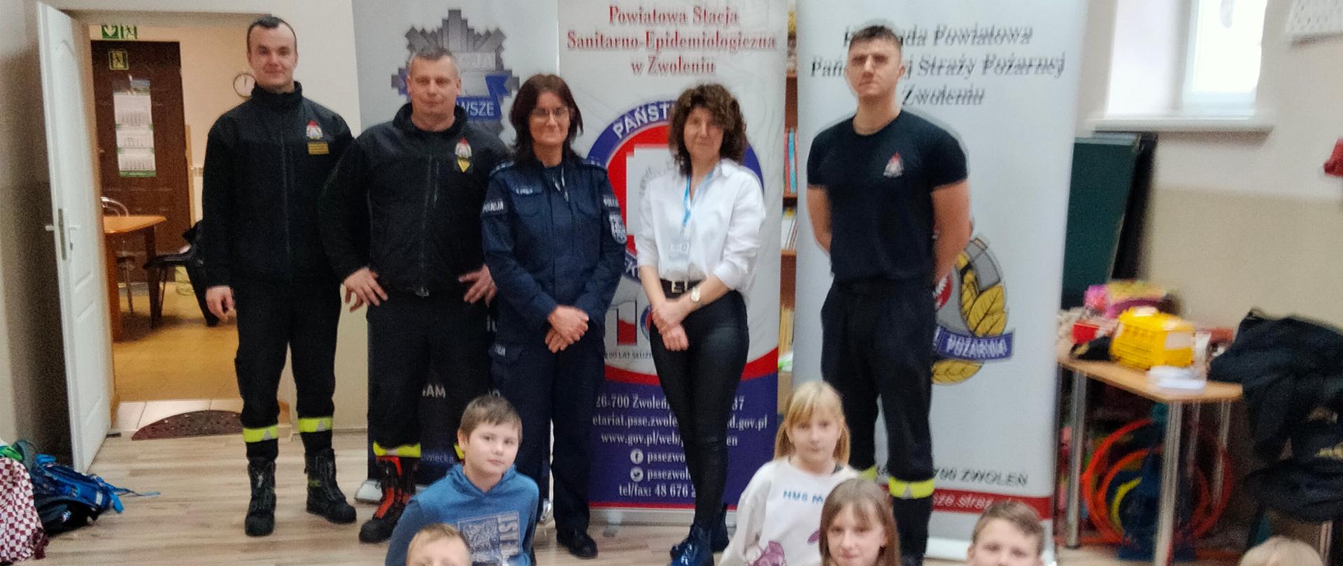 Zdjęcie przedstawia spotkanie edukacyjne, prawdopodobnie dotyczące pierwszej pomocy lub bezpieczeństwa, odbywające się w pomieszczeniu. Na pierwszym planie widać manekina do resuscytacji leżącego na dywanie, a wokół niego klęczą dzieci, z których jedno dotyka manekina. Za dziećmi i manekinem stoi grupa dorosłych, w tym strażacy w mundurach oraz cywile. W tle widoczne są dwa banery: jeden z napisem "Powiatowa Stacja Sanitarno-Epidemiologiczna w Zwoleniu" oraz drugi z logiem Państwowej Straży Pożarnej. Po prawej stronie widać stolik z akcesoriami ratowniczymi, takimi jak rękawiczki czy apteczka.