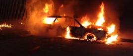 Płonące w całości auto na poboczu drogi przy ogrodzeniu. Pora nocna
