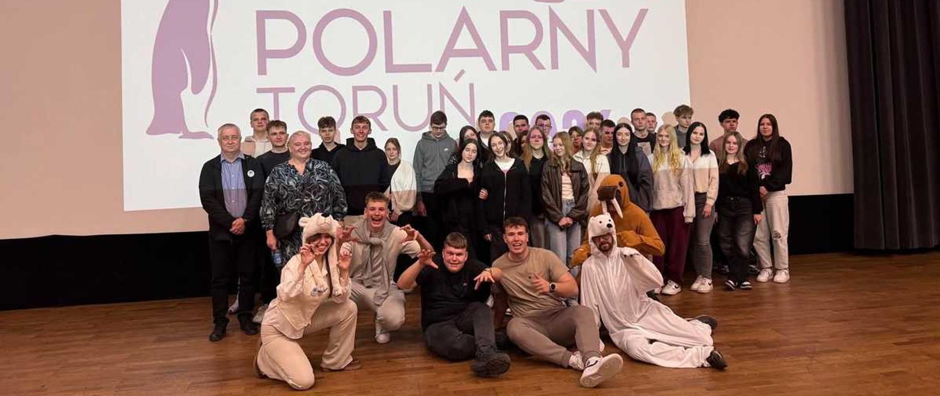 djęcie przedstawia grupę około 30 osób pozujących na scenie w auli wykładowej.Tło i ScenografiaGłównym elementem tła jest wielki ekran projekcyjny, na którym wyświetlany jest biały slajd z fioletowymi i różowymi akcentami. Napis na środku głosi: „DZIEŃ POLARNY TORUŃ”.Po lewej stronie napisu widnieje duża sylwetka pingwina, a nad napisem zarys niedźwiedzia polarnego.W lewym górnym rogu znajduje się logo Uniwersytetu Mikołaja Kopernika w Toruniu oraz Wydziału Nauk o Ziemi i Gospodarki Przestrzennej.Po prawej stronie ekranu zwisa ciężka, ciemna kotara. Podłoga wykonana jest z jasnobrązowych paneli.Osoby na zdjęciuGrupa składa się głównie z młodych ludzi (prawdopodobnie studentów) oraz kilku dorosłych.Pierwszy plan (siedzący/klęczący): Pięć osób siedzi bezpośrednio na podłodze. Dwie z nich mają na sobie białe, puszyste kostiumy zwierząt (prawdopodobnie niedźwiedzi polarnych) z kapturami z uszami. Pozostali są ubrani w stroje codzienne – bluzy i t-shirty.Drugi plan (stojący): Pozostała część grupy stoi w dwóch lub trzech rzędach. Panuje swobodna atmosfera – niektórzy uśmiechają się do aparatu, inni rozmawiają. Ubiór jest zróżnicowany: dominują bluzy z kapturem, swetry, dżinsy i ciemne kurtki.Wyróżnia się jedna osoba w głębi, która założyła maskę lub kostium morsa z charakterystycznymi ciosami.Atmosfera zdjęcia:Fotografia ma charakter pamiątkowy i radosny, dokumentując wydarzenie edukacyjne lub integracyjne związane z tematyką polarną na toruńskim uniwersytecie.