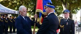 Wicewojewoda pomorski przekazuje sztandar komendantowi powiatowemu Państwowej Straży Pożarnej w Lęborku, obok stoi strażak z mikrofonem.