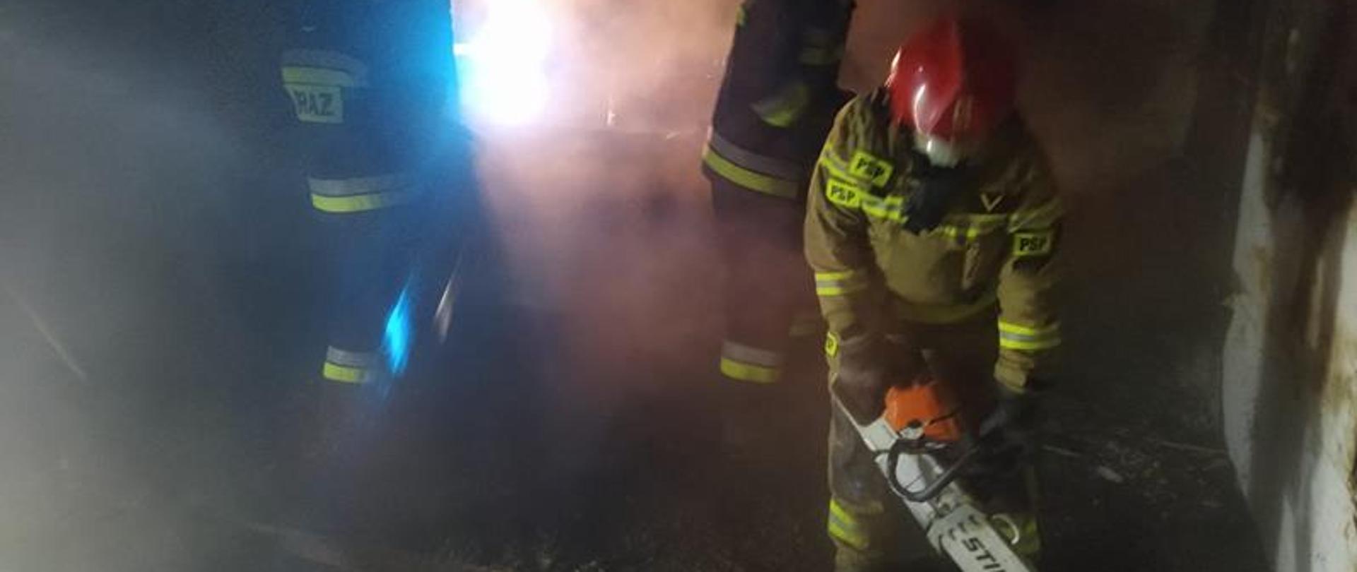  Trzech strażaków znajduje się w środku pomieszczenia w którym występował pożar. Jeden z nich wycina deski przy użyciu pilarki mechanicznej.