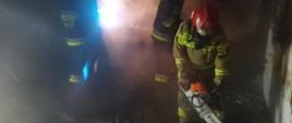  Trzech strażaków znajduje się w środku pomieszczenia w którym występował pożar. Jeden z nich wycina deski przy użyciu pilarki mechanicznej.