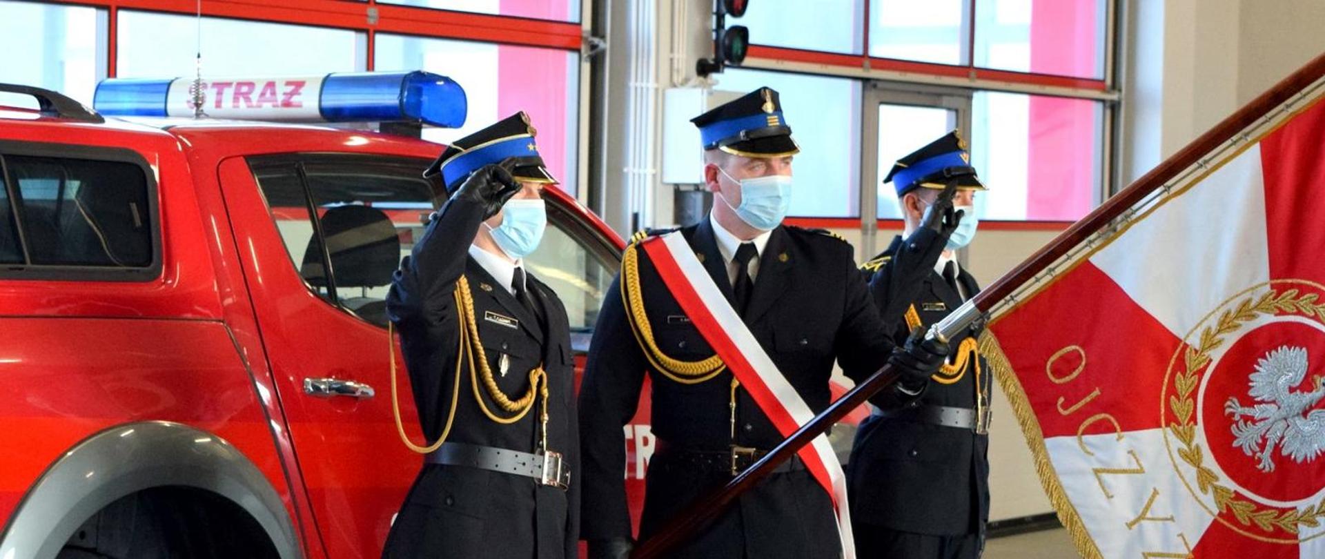 Zdjęcie zrobione w pomieszczeniu. Na zdjęciu widać poczet sztandarowy składający się z trzech strażaków, ubranych w mundury wyjściowe. Strażak ustawiony w środku przepasany jest biało-czerwona szarfą i trzyma pochylony sztandar. Obok niego funkcjonariusze salutują. Za strażakami widać samochód pożarniczy.