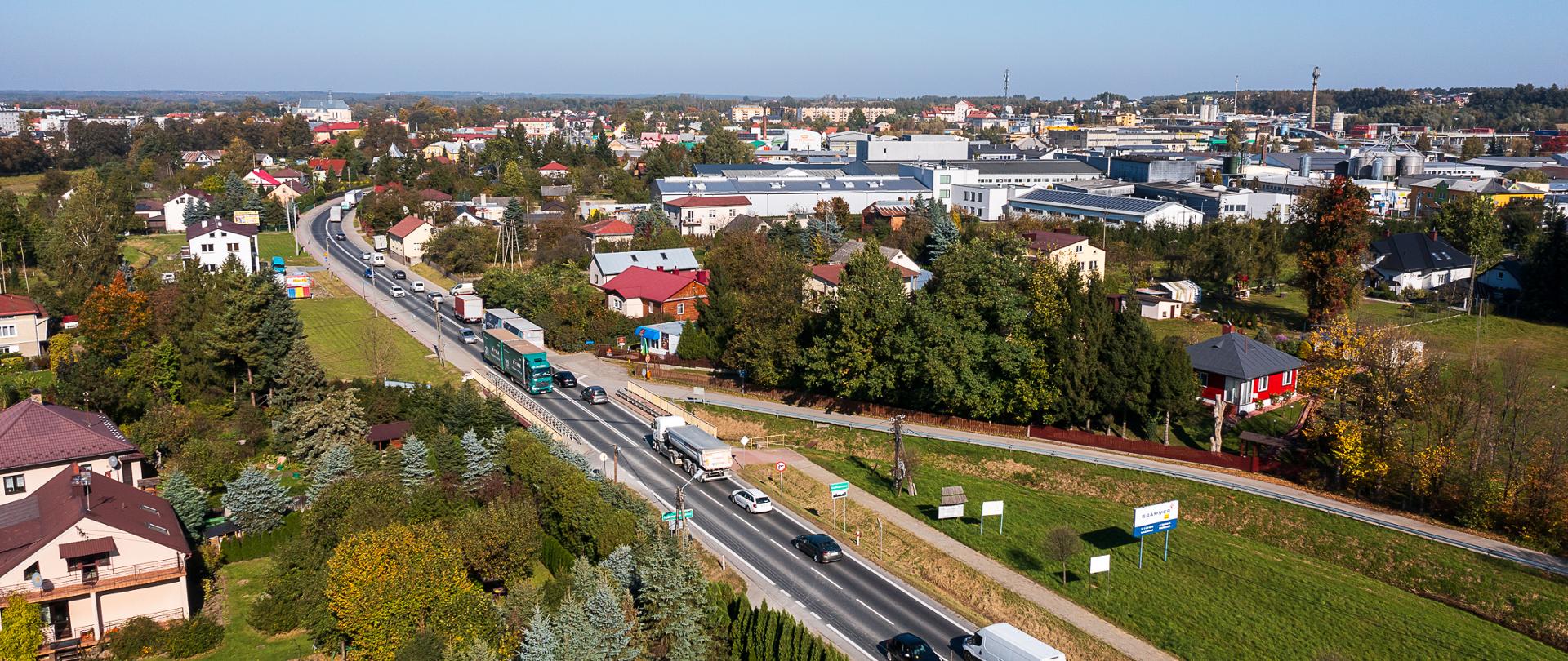 Widok z lotu ptaka na drogę krajowa nr 9 w miejscowości Kolbuszowa. Droga przebiega przez centrum miejscowości, po obu stronach drogi znajduje się ścisła zabudowa domów, na drodze duży ruch pojazdów osobowych i ciężarowych. Lato, świeci słońce. 