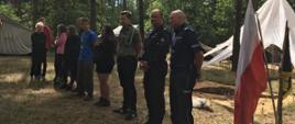Na zdjęciu wizytacja obozu harcerskiego przez służby mundurowe. Strażak i policjant przekazuje informacje o zagrożeniach na obozowisku, harcerze stoją w lesie w ubraniach cywilnych w tle widać namioty oraz flagę biało-czerwoną.