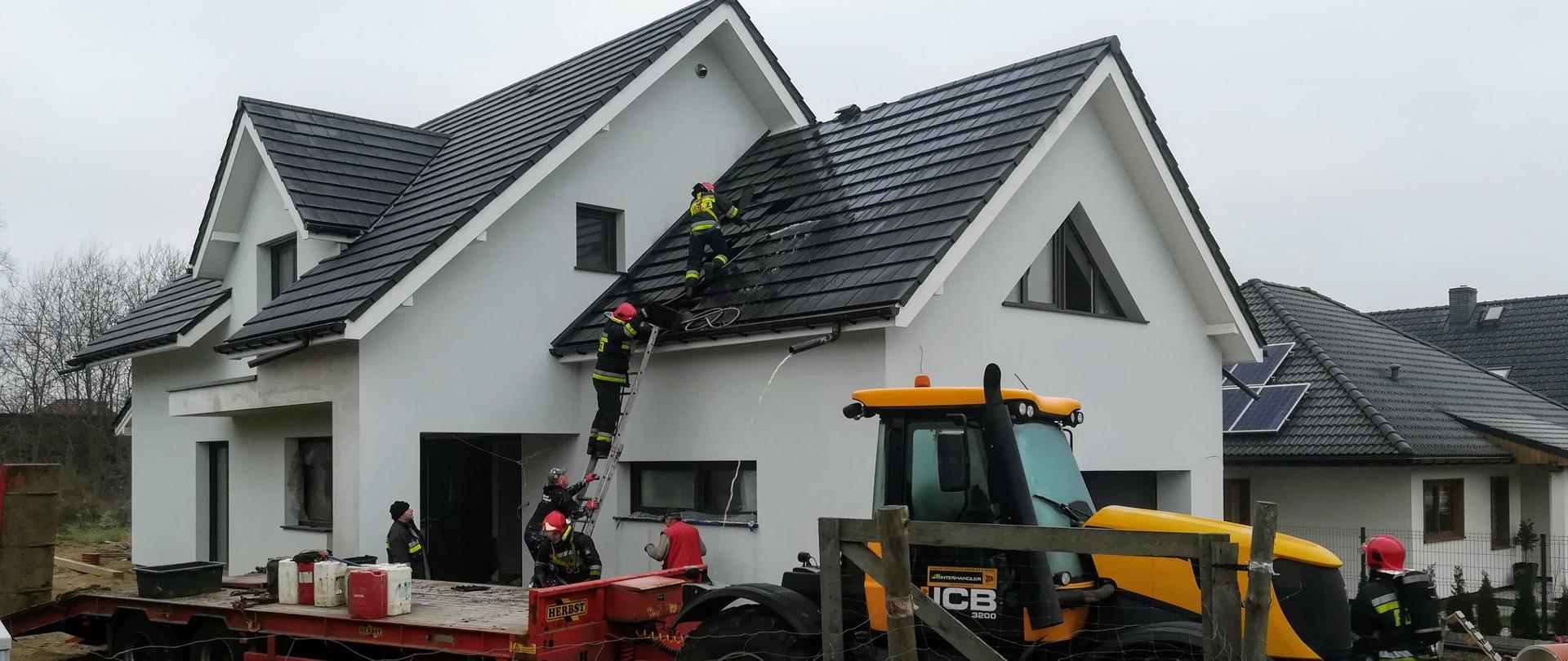 Dom jednorodzinny, na podwórku traktor, na dach budynku wchodzą po drabinie strażacy