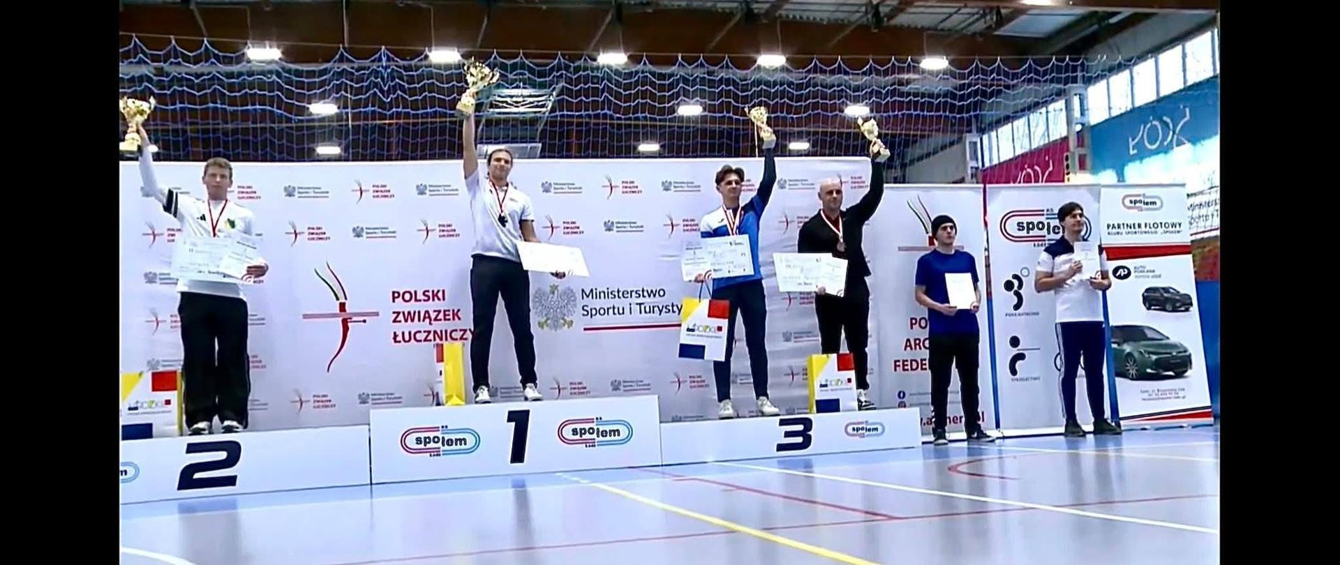 sportowcy stoją na podium, podnoszą do góry puchary