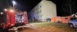Widok na budynek wielorodzinny. Po lewej stronie widoczny samochód gaśniczy sochaczewskiej straży pożarnej, a po lewej namiot ratowniczy w kolorze pomarańczowym oraz radiowóz policji.