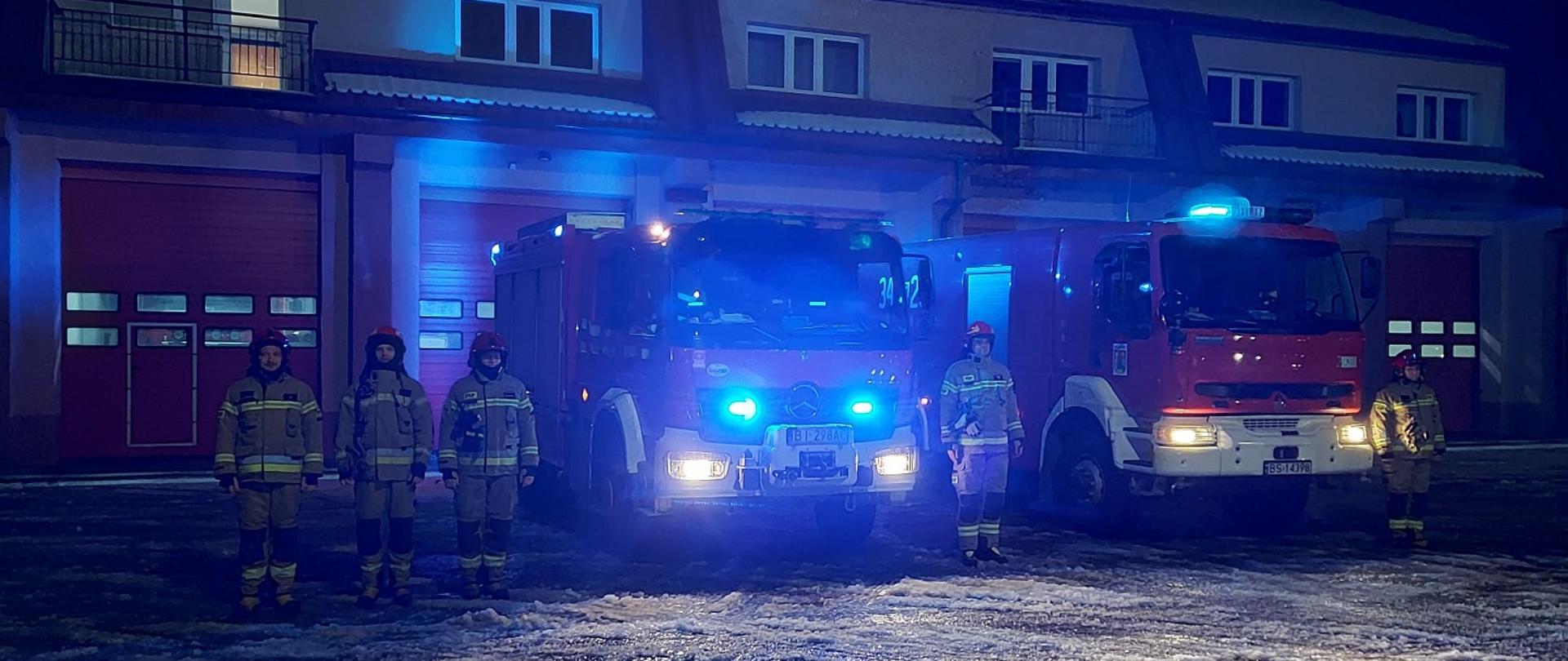 Zdjęcie obrazuje strażaków, samochody pożarnicze w tle budynek w porze nocnej.