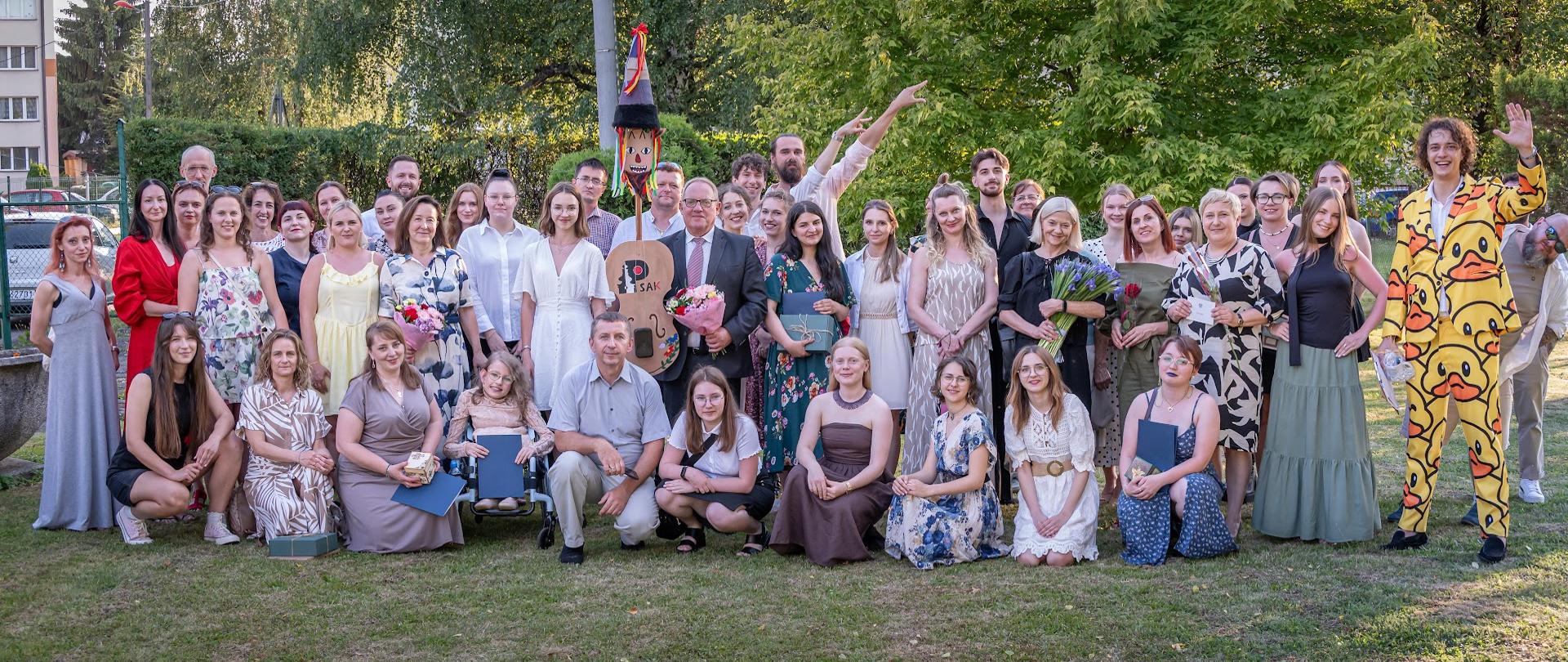 Zdjęcie przedstawia dużą grupę słuchaczy i nauczycieli podczas zakończenia roku szkolnego. Wszyscy pozują w grupie na tle trawy i zielonych drzew. 