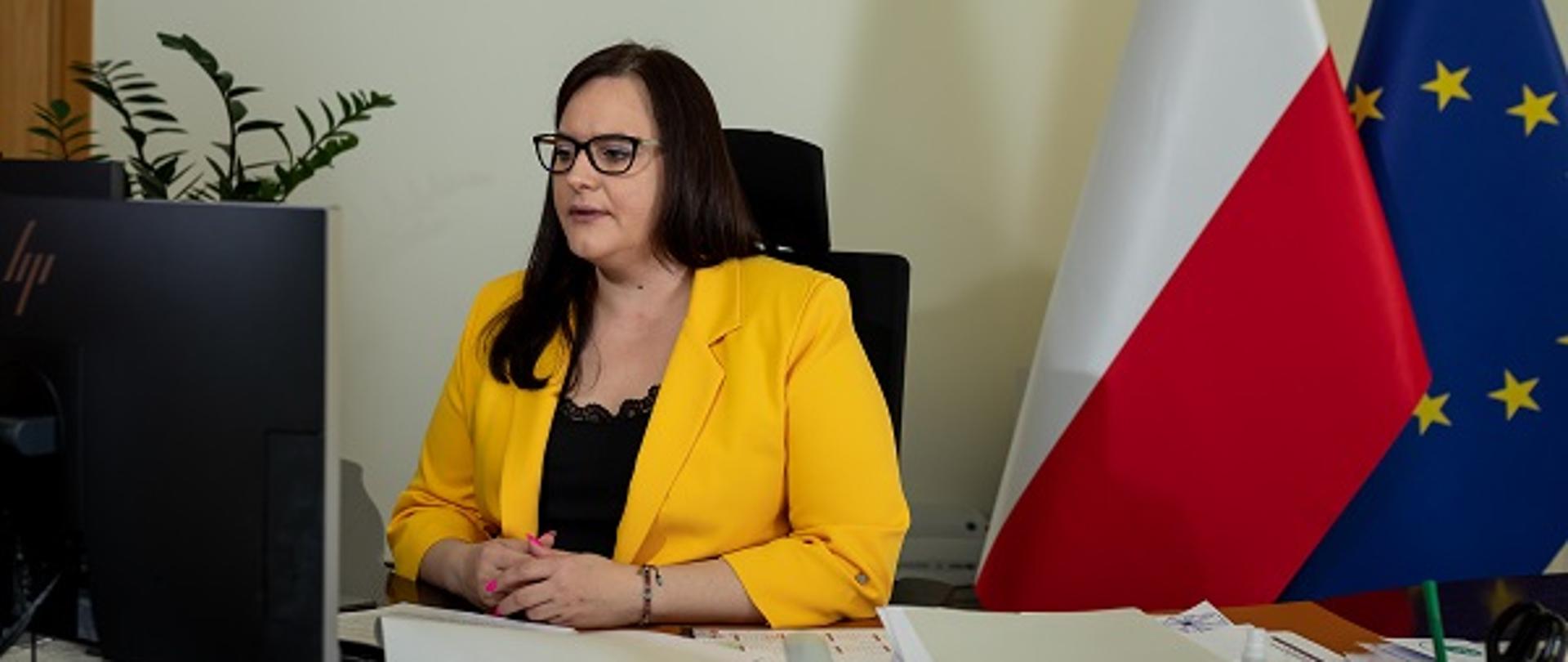 Minister Małgorzata Jarosińska-Jedynak siedzi za biurkiem przed komputerem, za plecami flagi UE i Polski