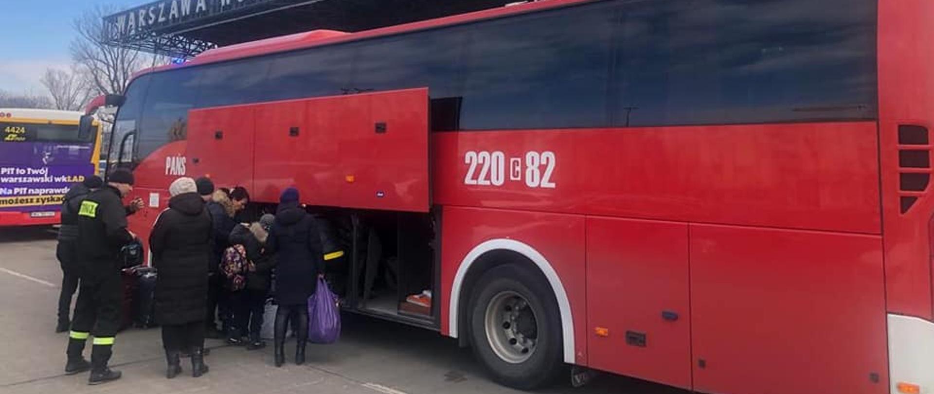 Zdjęcie przedstawia czerwony autobus stojący na utwardzonym placu. Przy otwartym bocznym luku bagażowym stoją ludzie i wkładają do wewnątrz walizki i torby.
