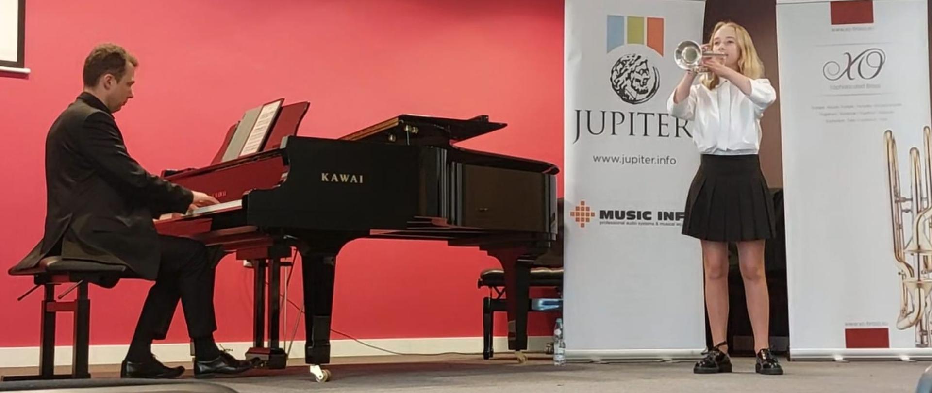 Zdjęcie numer 2 podczas występu na tle dwóch białych rolapów z napisem Jupiter Music info oraz drugiego niezidentyfikowanego z fragmentem złotego instrumentu dętego uczennica Patrycja szałapata wykonuje na trąbce utwór przy fortepianie marki kałaj nauczyciel Dominik Zabłocki
