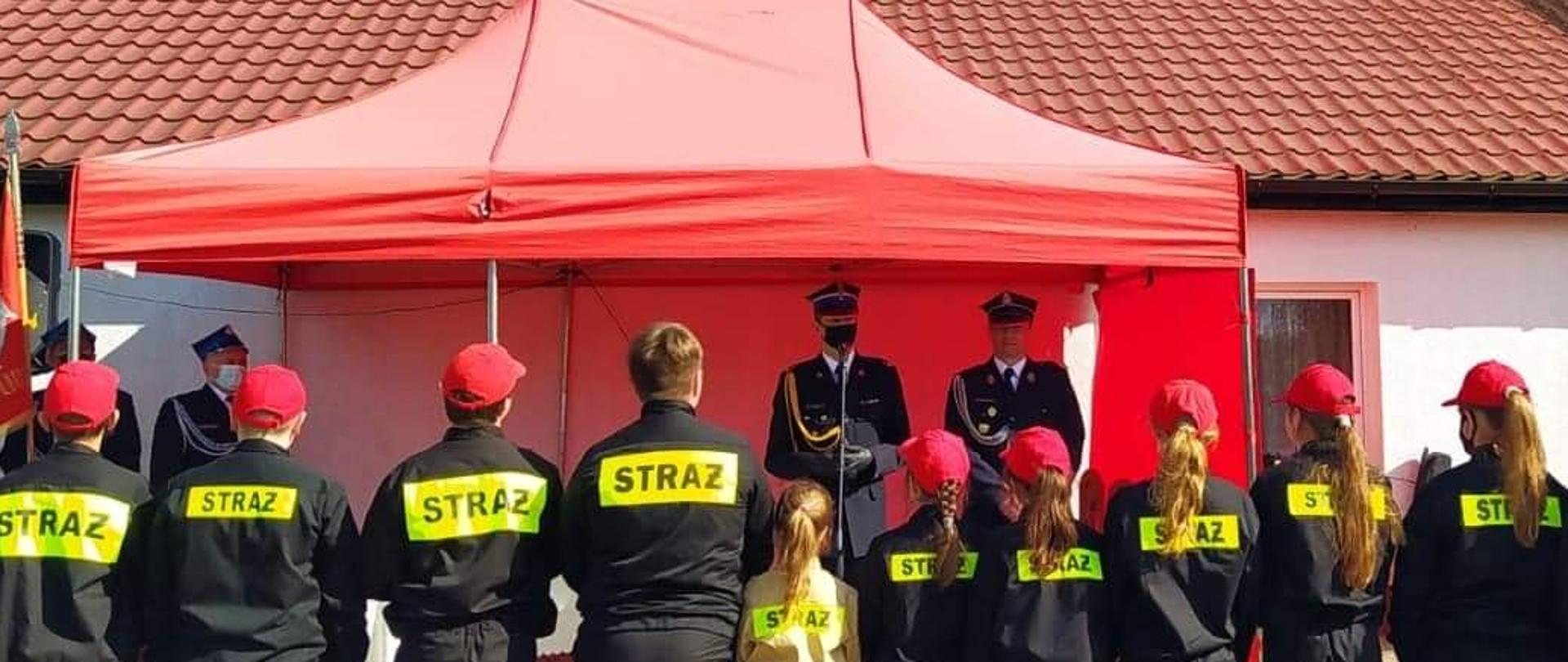 Na zdjęciu dzieci z młodzieżowej drużyny pożarniczej stojące na baczność na wprost przemawiającego Komendanta. Wszyscy w umundurowaniu strażackim.