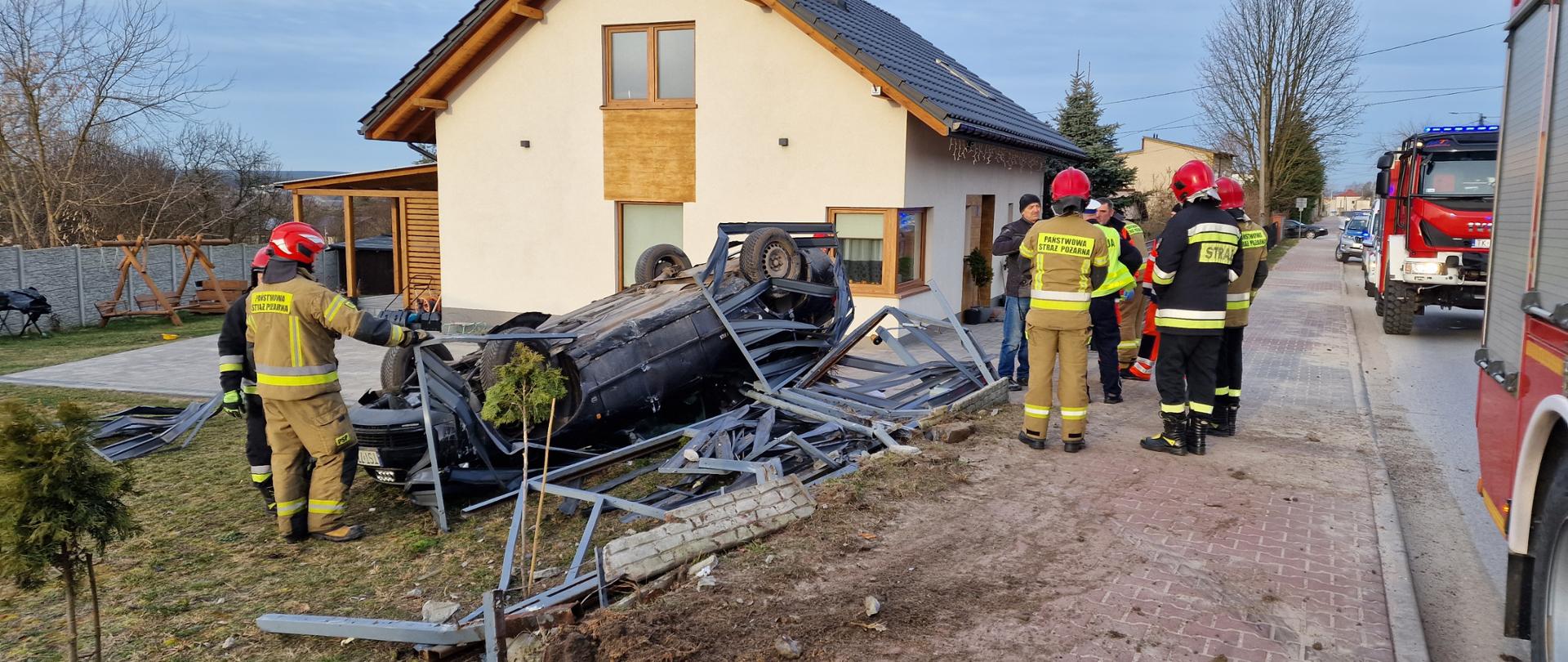 Samochód znajdujący się na dachu , zniszczone ogrodzenie. obok stoją strażacy
