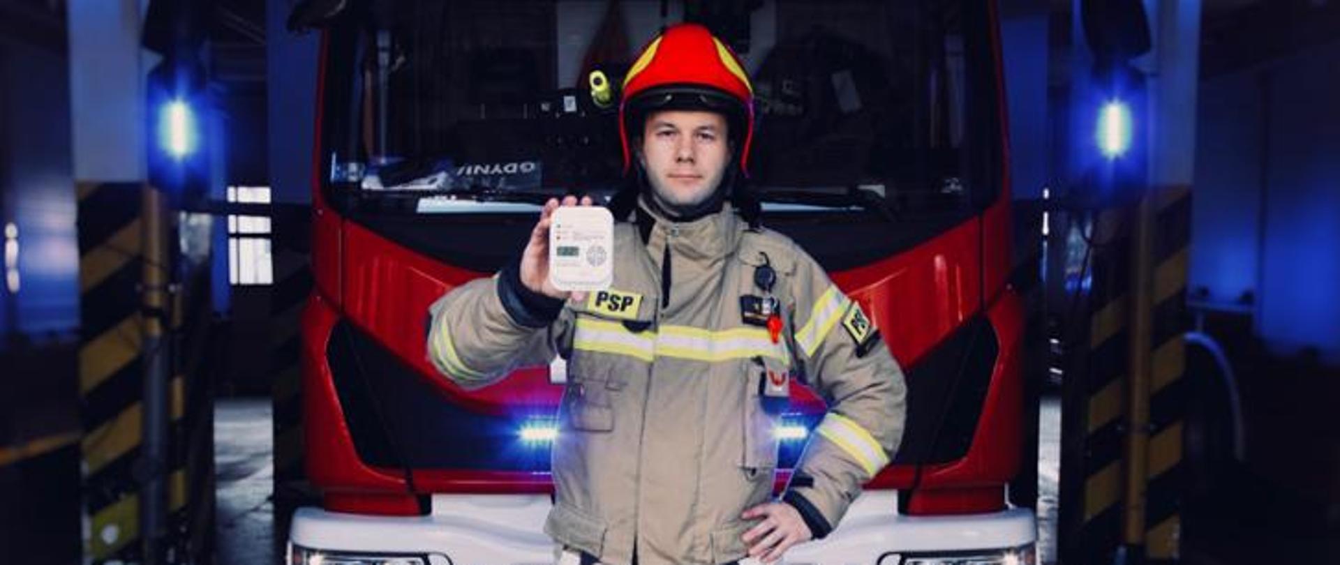 Na grafice strażak ubrany w strój bojowy trzyma w ręku czujkę pożarową. Za strażakiem znajduje się wóz strażacki.