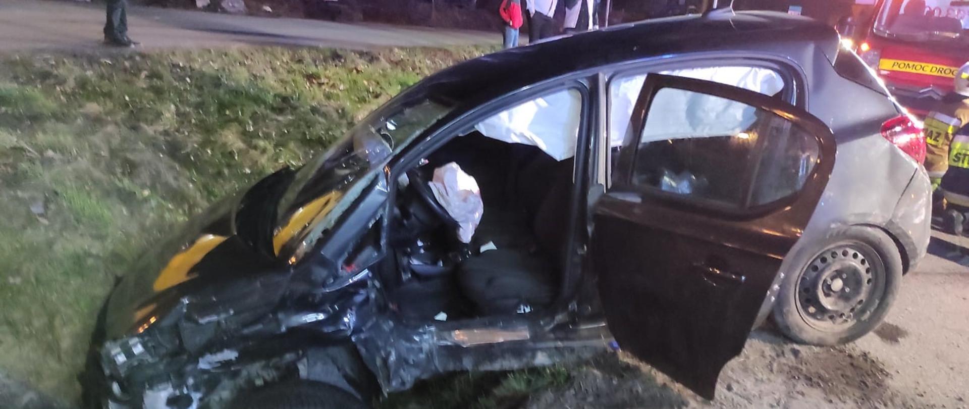 Na zdjęciu widoczny samochód osobowy znajdujący się w przydrożnym rowie. Lewa strona pojazdu zniszczona w wyniku uderzenia innego pojazdu oraz działania służb ratowniczych, które musiały usunąć drzwi w celu wykonania dostępu do poszkodowanych osób znajdujących się wewnątrz. Z prawej strony częściowo widoczny pojazd Pomocy drogowej i strażacy udzielający pomocy poszkodowanym. Na drugim planie powyżej drogi krajowej widoczne drzewa oraz osoby postronne przyglądające się prowadzonej akcji ratowniczej.