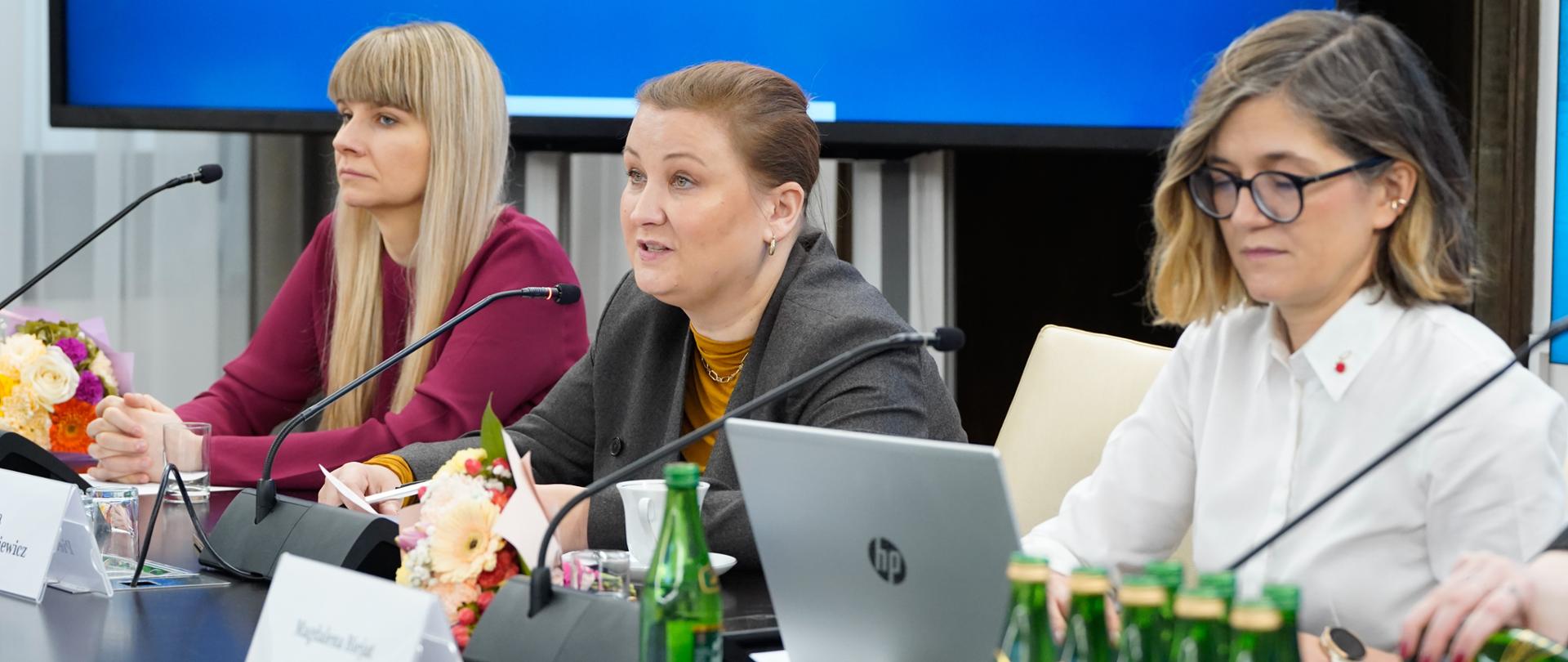 Na zdjęciu widać siedzące przy stole prezydialnym (od lewej): Rzecznik Praw Dziecka Monikę Horną-Cieślak, wiceminister w MEN Paulinę Piechnę-Więckiewicz oraz wicemarszałek Senatu Magdalenę Biejat. W trakcie wykonywania zdjęcia wiceminister Paulina Piechna-Więckiewicz przemawia do włączonego mikrofonu. Na stole widać również szklanki, butelki z wodą, identyfikatory imienne, pozostałe mikrofony oraz bukiety kwiatów. W tle widać ekran z napisem "Senat Rzeczypospolitej Polskiej".