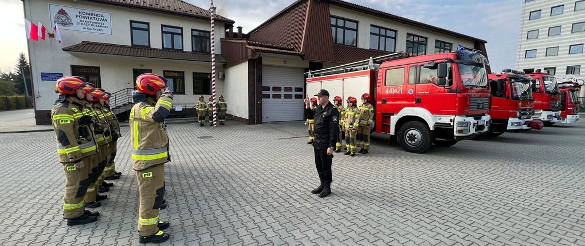 Na zdjęciu widoczny na placu wewnętrznym przed Komendą PSP w Gorlicach poczet flagowy przy maszcie w ubraniach specjalnych w kolorze piaskowym i czerwonych hełmach podczas podnoszenia flagi w zawiązku z uroczystością Dnia Flagi Rzeczypospolitej Polskiej. Przed pocztem ustawione zmiany służbowe w dwuszeregu w pozycji na baczność również w ubraniach specjalnych koloru pisakowego i czerwonych hełmach. 