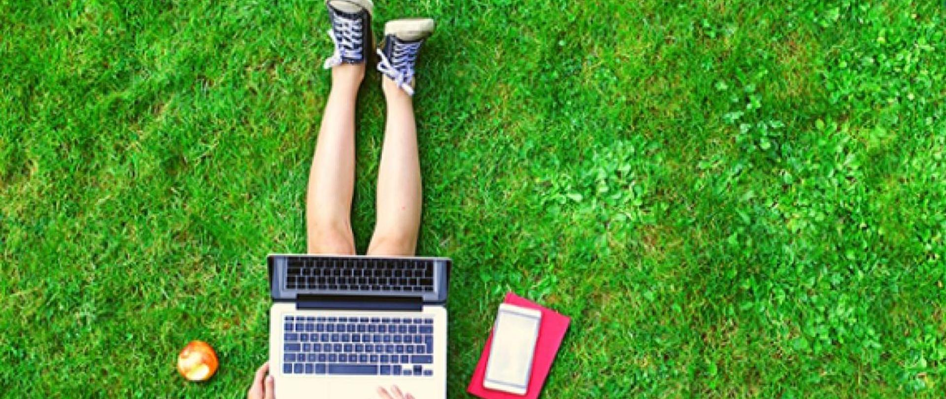 dziecko siedzi z laptopem na trawie. obok jabłko i notatnik