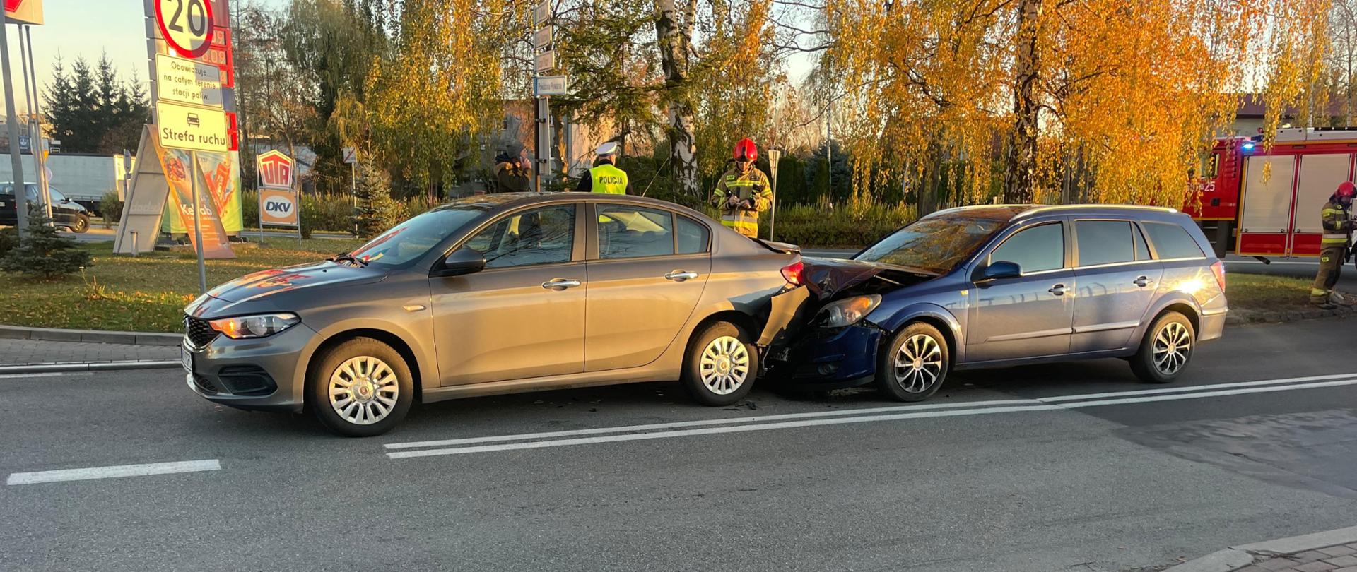 Na zdjęciu widać dwa samochody osobowe po kolizji, stoją na drodze: szary sedan z przodu i niebieskie kombi z tyłu, z widocznymi uszkodzeniami zderzaków. W tle, na poboczu, widać policjantów i strażaków oraz fragmenty wozów służbowych, a także drzewa z jesiennym listowiem. Kolizja miała miejsce w pobliżu stacji paliw, widoczne pylony z cenami paliw.