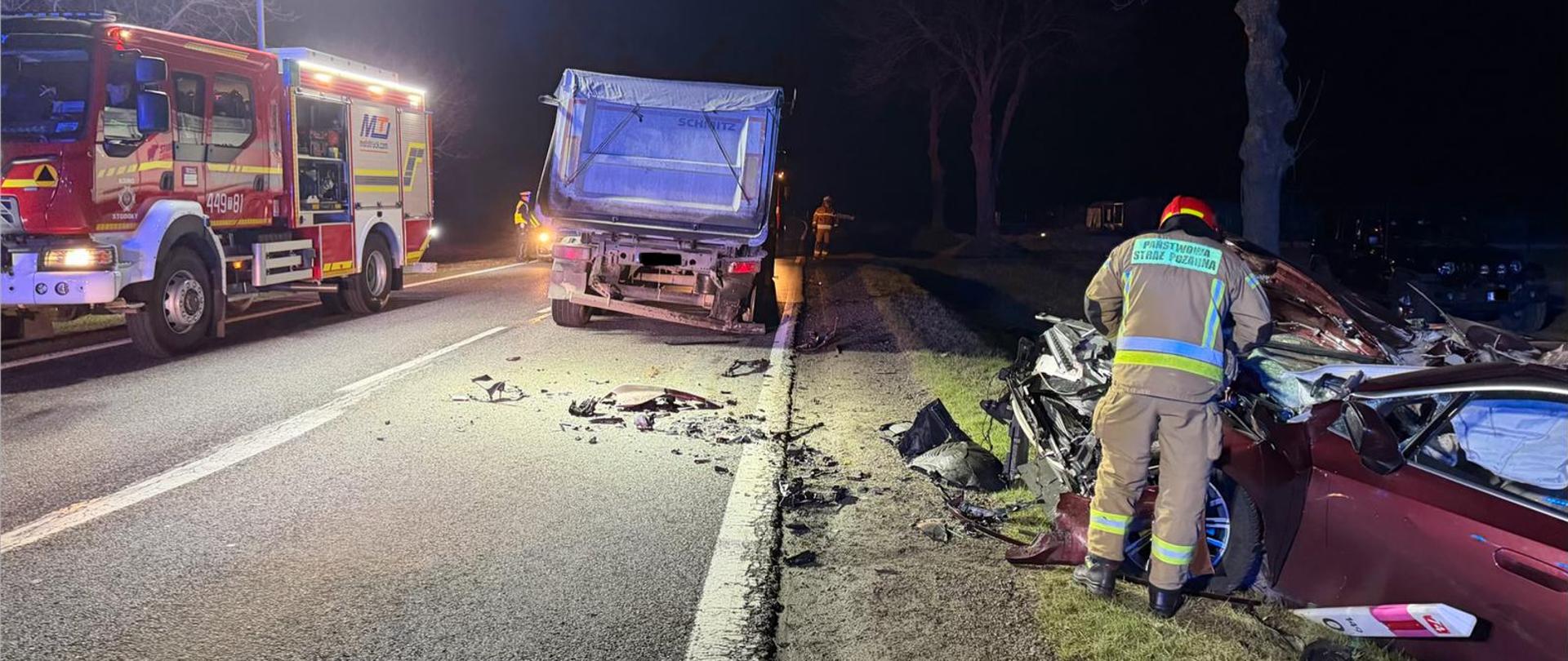 Nocna scena wypadku drogowego na nieoświetlonej drodze. Na poboczu stoi poważnie uszkodzony czerwony samochód osobowy, przy którym strażak w odblaskowym stroju prowadzi działania ratunkowe. Na jezdni widać rozrzucone fragmenty pojazdów. Przed autem stoi ciężarówka. Po lewej stronie zaparkowany jest wóz strażacki z włączonymi światłami, oświetlający miejsce zdarzenia. W tle widoczne są drzewa i kilku ratowników pracujących przy zabezpieczaniu miejsca wypadku