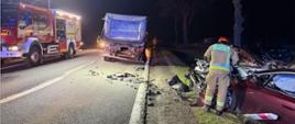 Nocna scena wypadku drogowego na nieoświetlonej drodze. Na poboczu stoi poważnie uszkodzony czerwony samochód osobowy, przy którym strażak w odblaskowym stroju prowadzi działania ratunkowe. Na jezdni widać rozrzucone fragmenty pojazdów. Przed autem stoi ciężarówka. Po lewej stronie zaparkowany jest wóz strażacki z włączonymi światłami, oświetlający miejsce zdarzenia. W tle widoczne są drzewa i kilku ratowników pracujących przy zabezpieczaniu miejsca wypadku