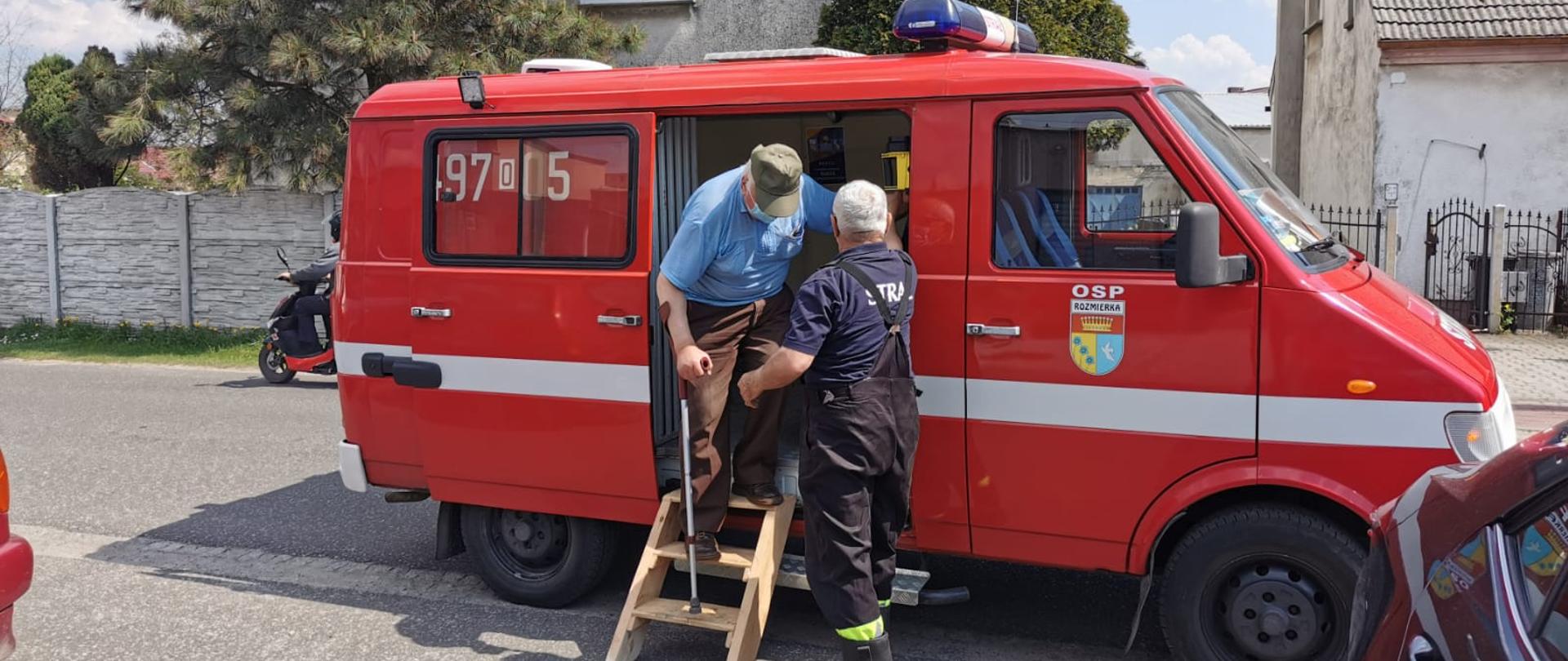 Zdjęcie przedstawia druha OSP Rozmierka, który pomaga wysiąść osobie, która została przetransportowana do punktu szczepień.