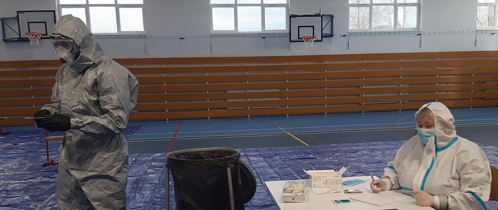 Fotografia została zrobiona na szkolnej sali gimnastycznej. W tle widać ścianę, na której zawieszone są dwie tablice do gry w koszykówkę oraz okna, poniżej których znajdują się kaloryfery zabezpieczone drewnianymi deskami. Na podłodze została rozłożona niebieska folia. W lewej części fotografii widzimy strażaka ubranego w szczelny szary kostium, na jego twarzy znajdują się duże okulary ochronne oraz maska medyczna a czarne rękawiczki przykrywają dłonie, w których trzyma pałeczki do wymazu. W centralnej części fotografii znajduję się duży kosz na odpady. Po prawej stronie można zobaczyć kobietę siedzącą za stołem, ona również ubrana jest w kombinezon ochronny oraz maseczkę i rękawiczki. Na stole znajdują się wymazówki, środki ochronne, dokumenty oraz taca z testami.