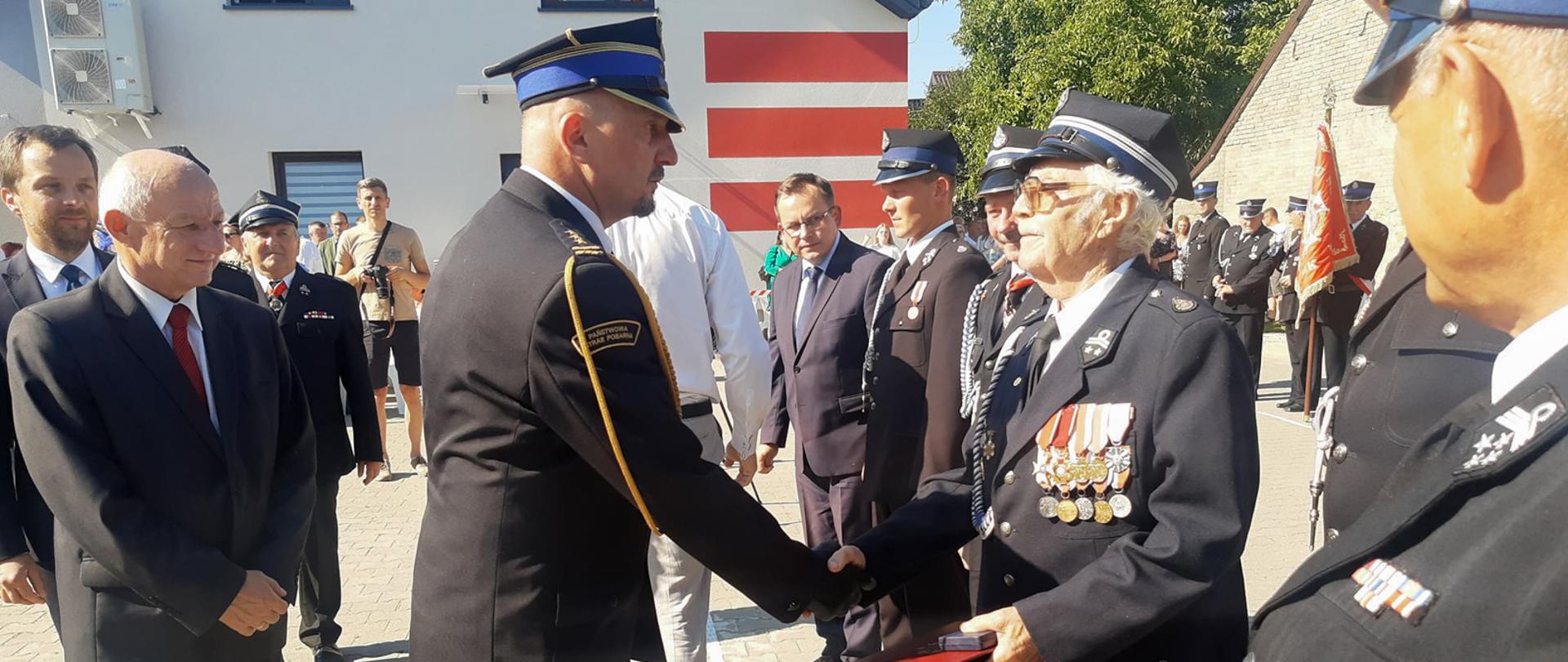 st. bryg. Paweł Malinowski gratuluje odznaczenia druhowi seniorowi OSP, na zdjęciu widać ludzi i druhów stojących na placu w czasie uroczystości