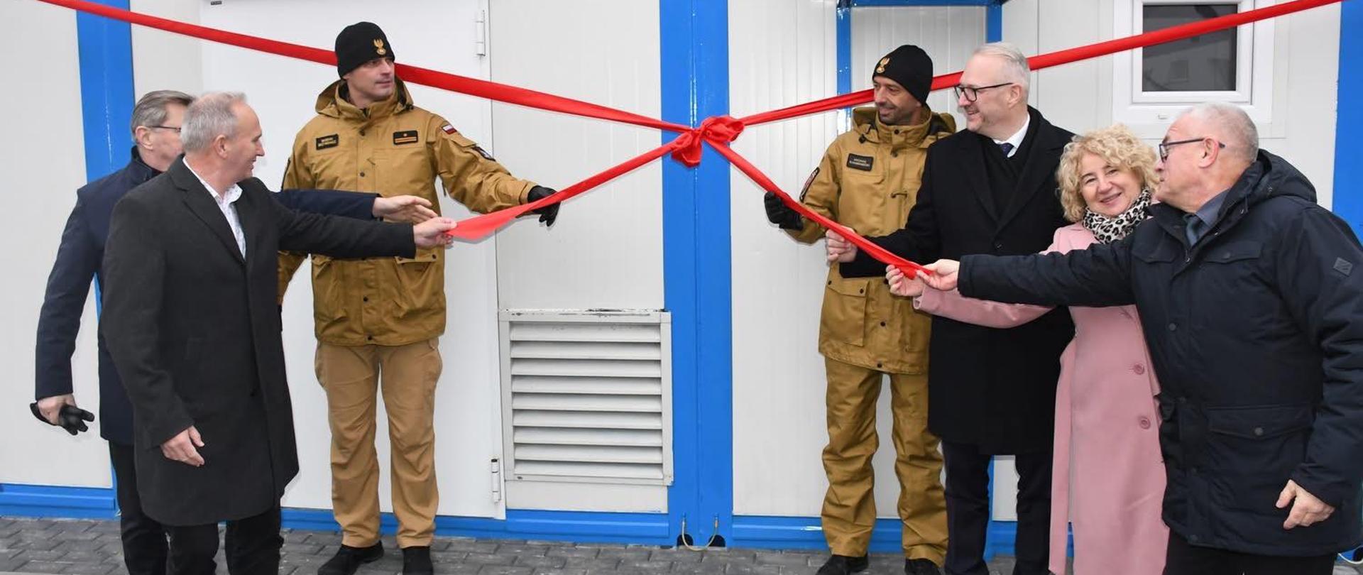 Na zdjęciu widoczna jest uroczystość przecięcia wstęgi przed nowym budynkiem modułowym. Siedem osób stoi w rzędzie na utwardzonym podłożu z kostki brukowej i asfaltu. Wszyscy trzymają rozciągniętą czerwoną wstęgę, która ma duży dekoracyjny węzeł pośrodku. Dwie osoby po lewej stronie ubrane są w beżowe mundury. Pozostałe osoby mają na sobie eleganckie płaszcze i kurtki.