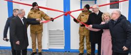 Na zdjęciu widoczna jest uroczystość przecięcia wstęgi przed nowym budynkiem modułowym. Siedem osób stoi w rzędzie na utwardzonym podłożu z kostki brukowej i asfaltu. Wszyscy trzymają rozciągniętą czerwoną wstęgę, która ma duży dekoracyjny węzeł pośrodku. Dwie osoby po lewej stronie ubrane są w beżowe mundury. Pozostałe osoby mają na sobie eleganckie płaszcze i kurtki.